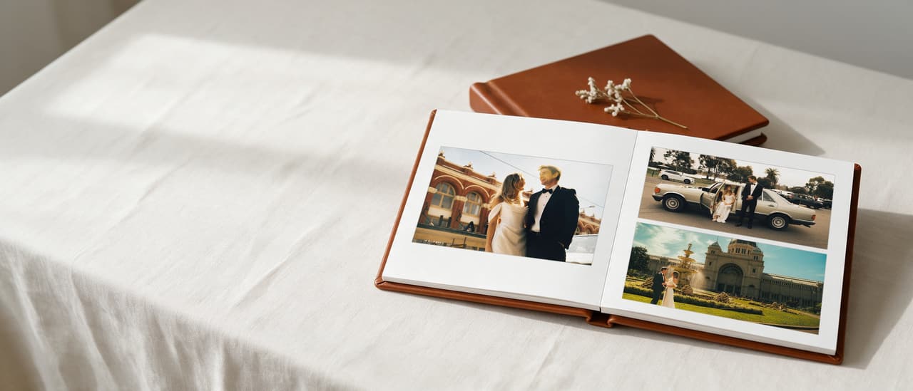 An open cognac leather layflat album: a couple laughing beside a heritage building on the left page, the bride in a classic car and the couple by a fountain on the right