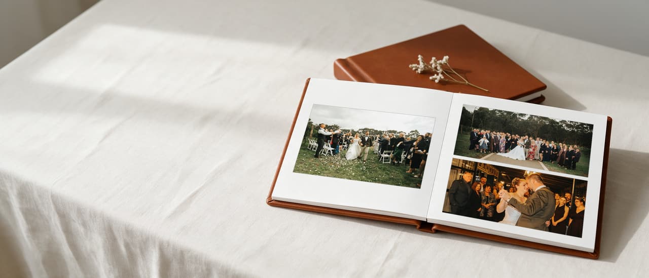 An open cognac leather layflat album on a linen tablecloth: a couple leaving their vineyard ceremony through falling petals on the left page, all their guests together and their first dance on the right, a second album behind it