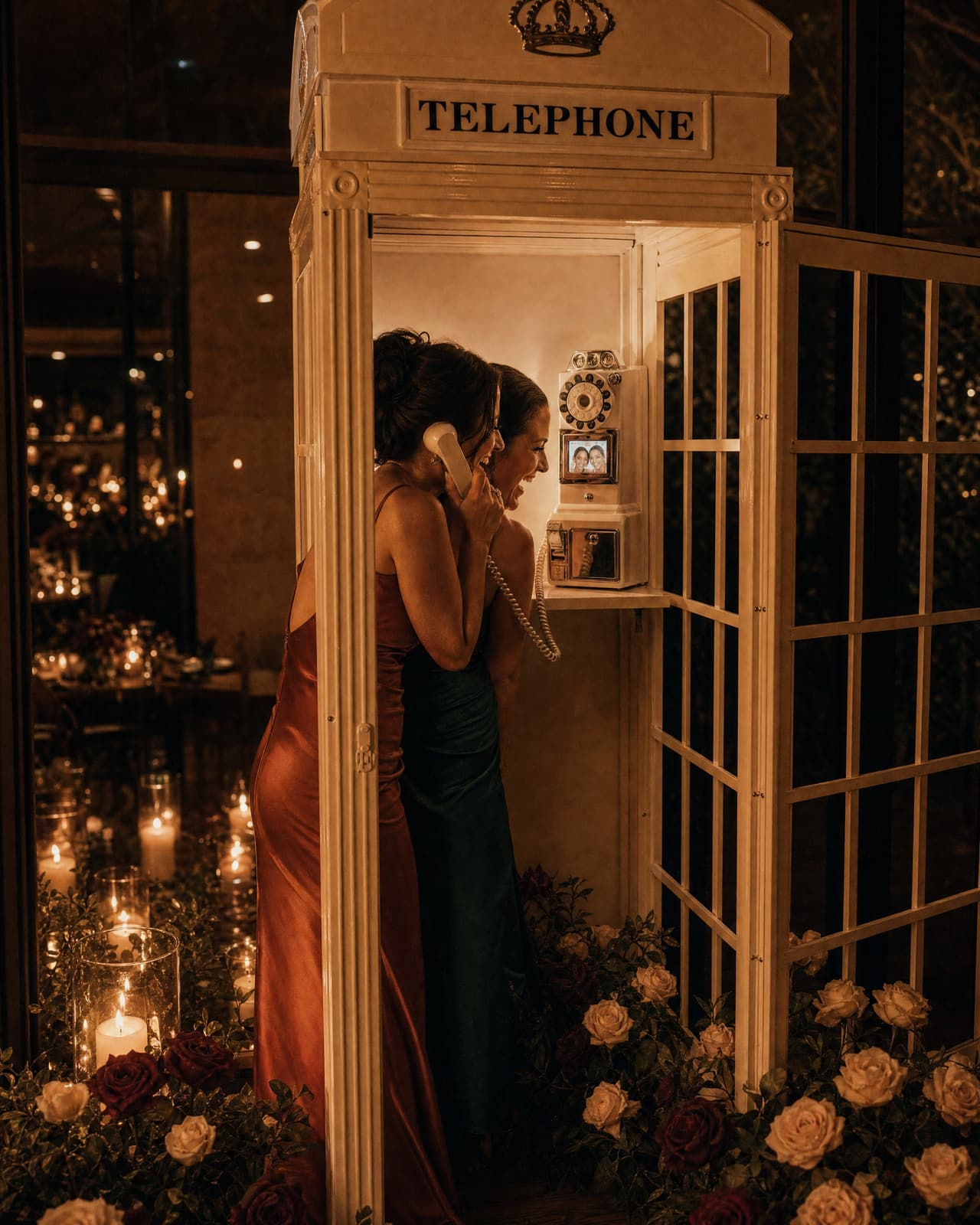 Two friends in rust and emerald dresses leaning in to the video phone inside the telephone booth at night, laughing at their own faces on its screen