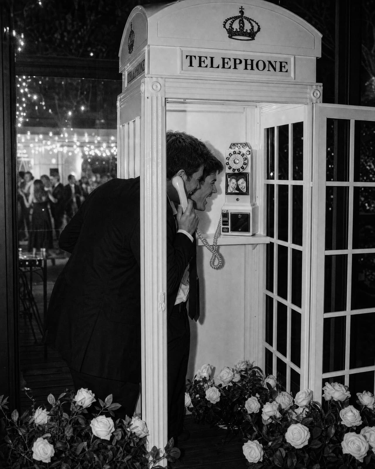 Black and white photo of two groomsmen leaning into the telephone booth late at night, laughing into the screen of the video phone as they record a message