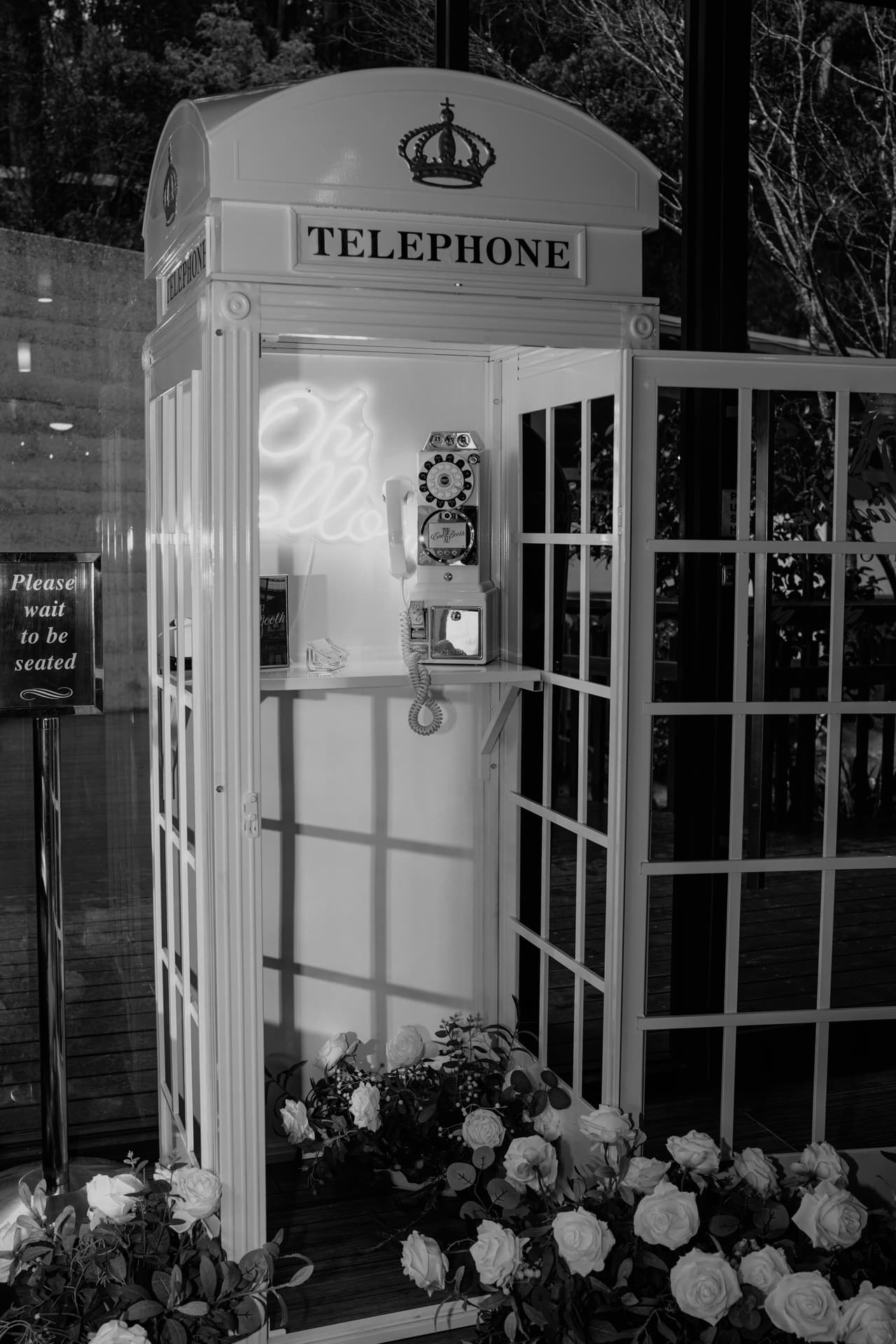 A white vintage telephone booth with its door open, roses and greenery around its base