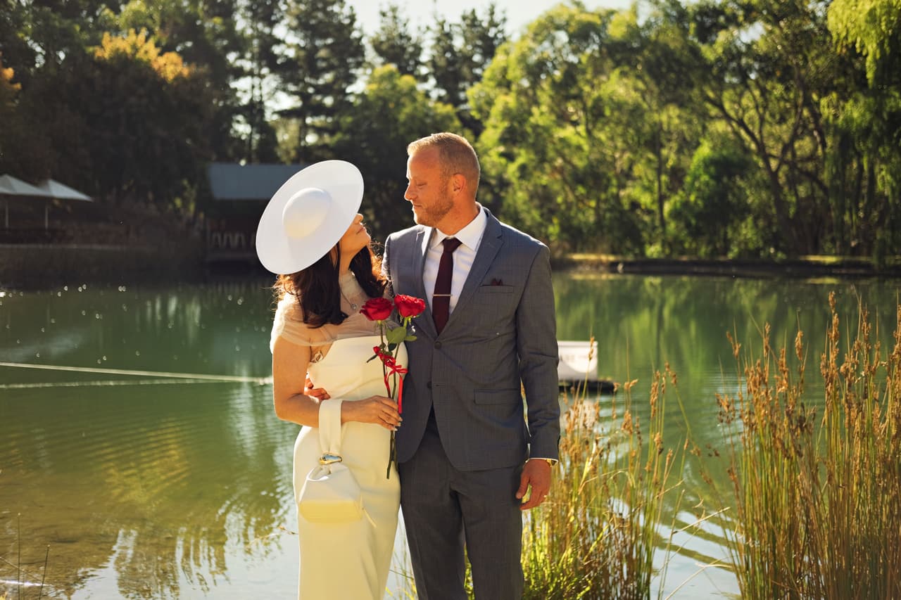 Kim in her wide hat looking up at Ben on the bank of the pond, the Boatshed across the water behind them