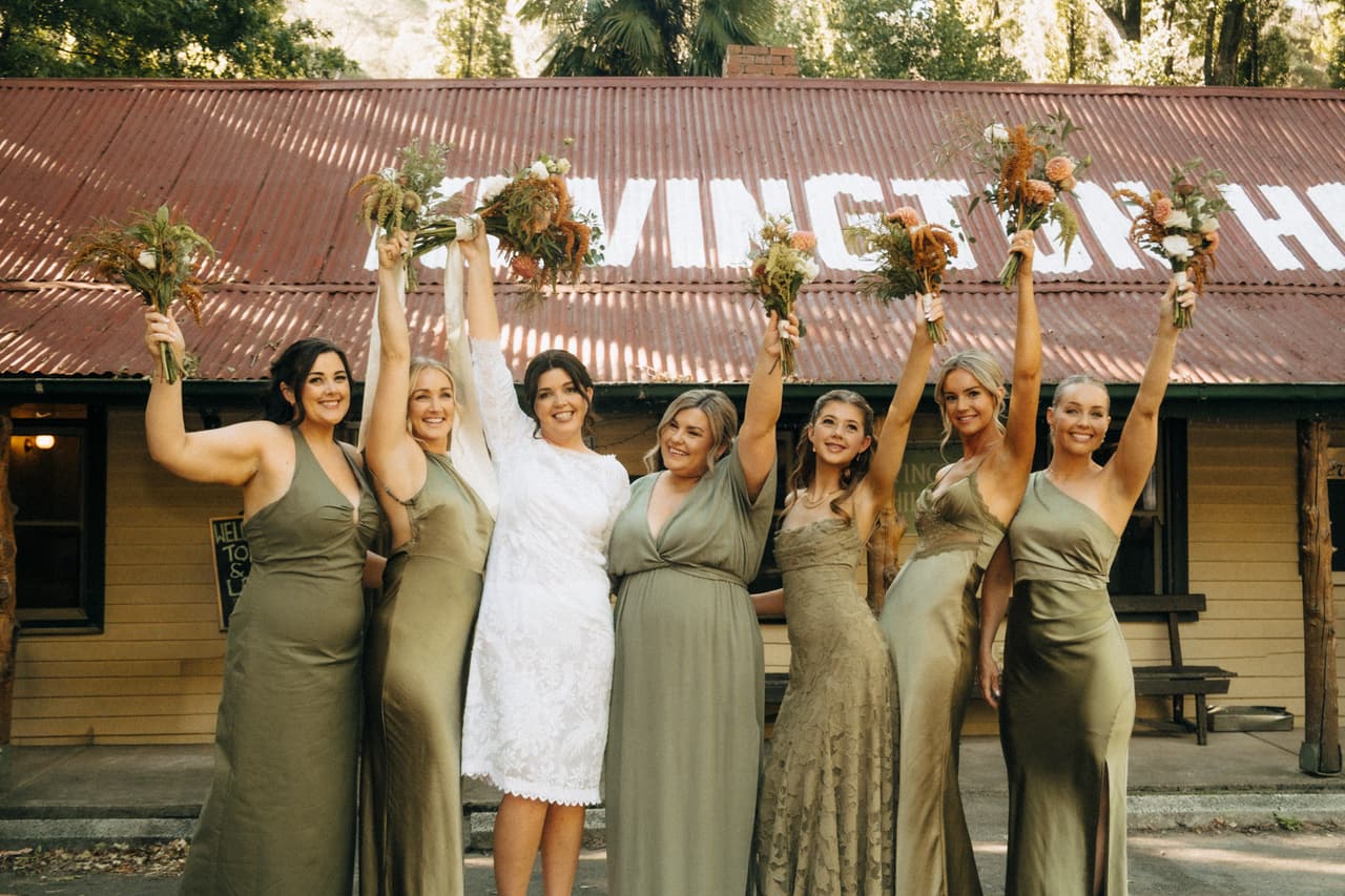 Bridesmaids in olive dresses and the bride raising their bouquets outside the Kevington Hotel