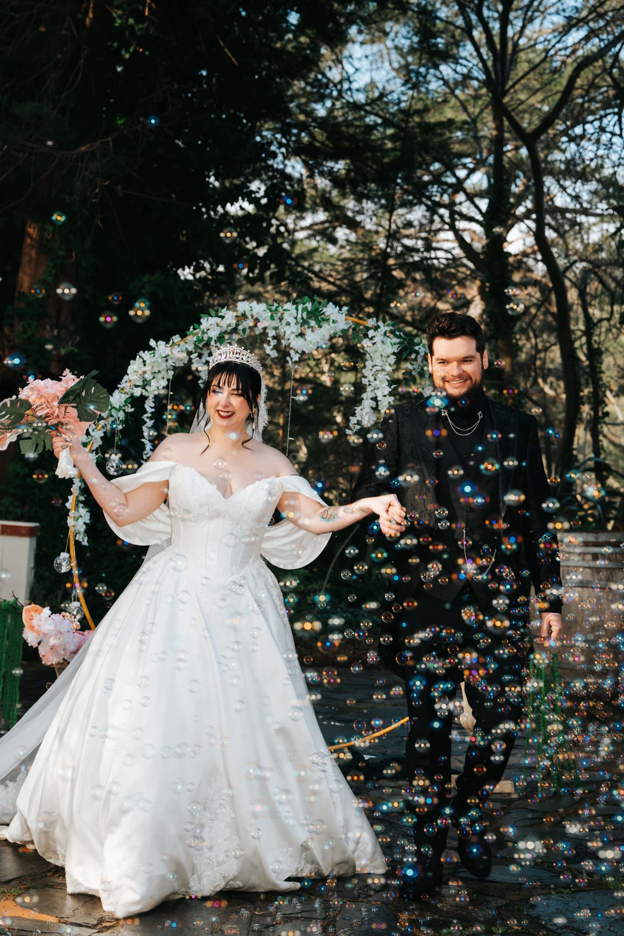 A bride laughing as she and the groom walk through a cloud of bubbles under a floral arch
