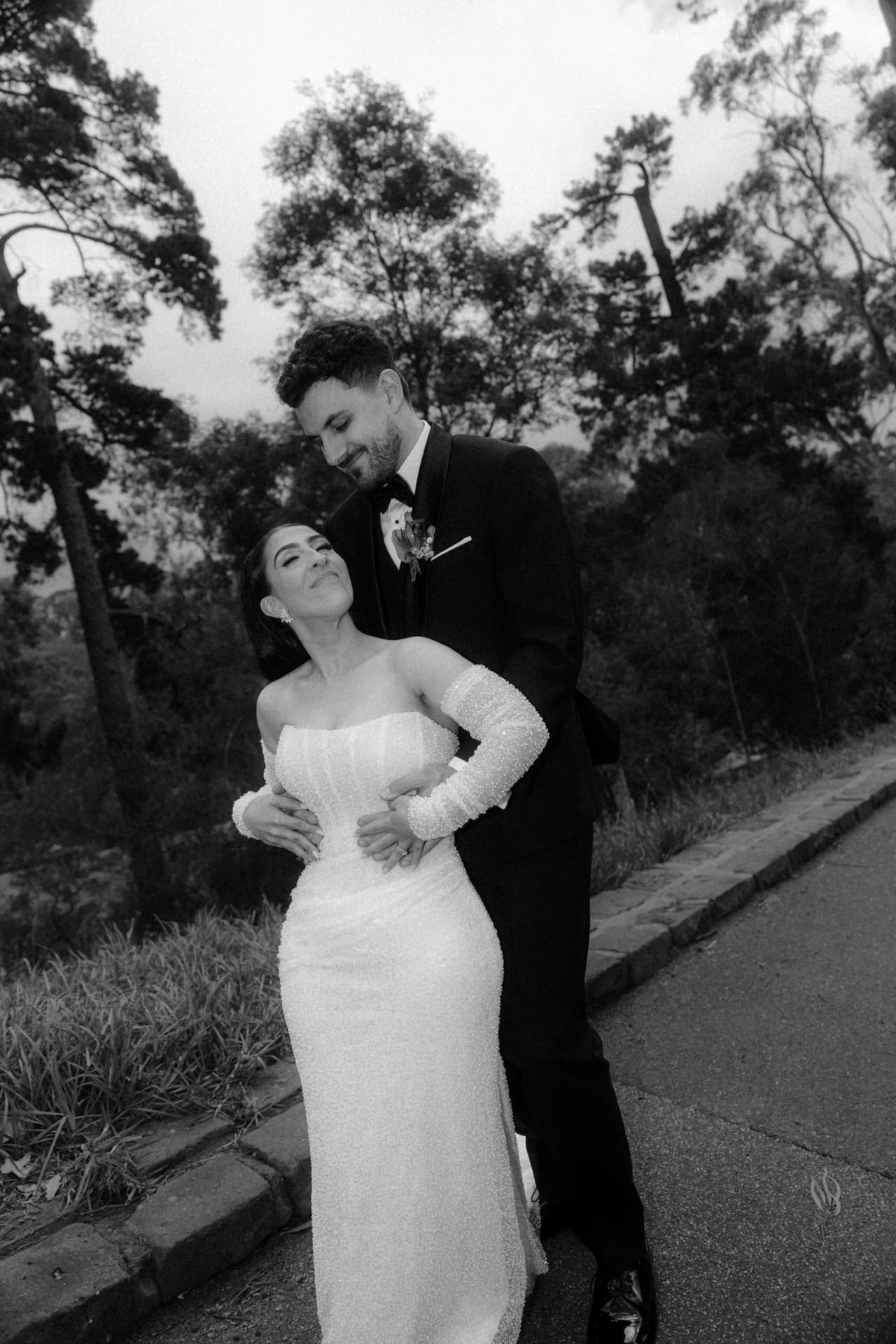 Black and white photo of a groom holding the bride from behind as she leans back laughing, beside a road through the trees