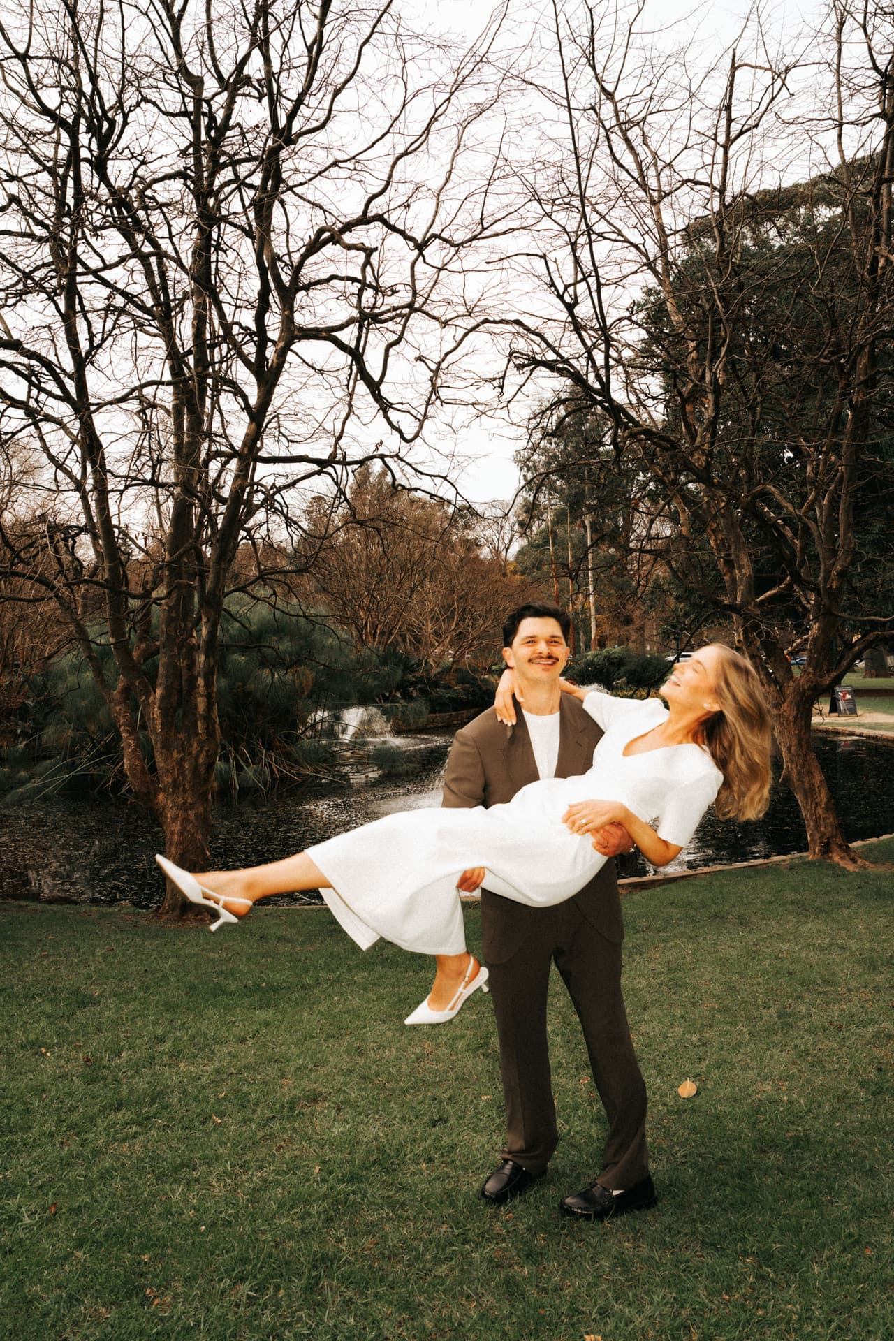 A groom carrying the bride in his arms across a lawn among bare winter trees