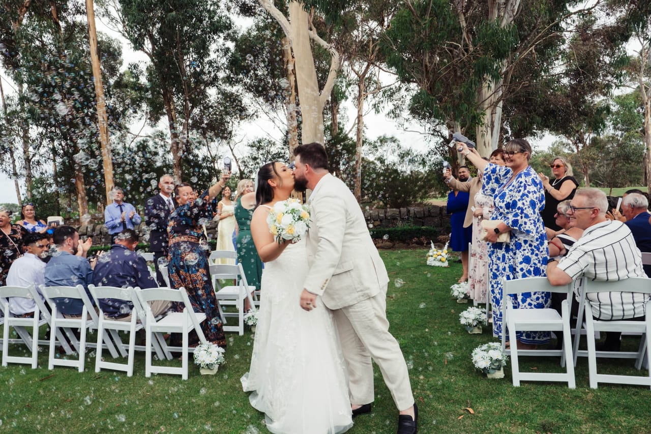 A couple kissing as they walk back down the aisle between cheering guests in a garden