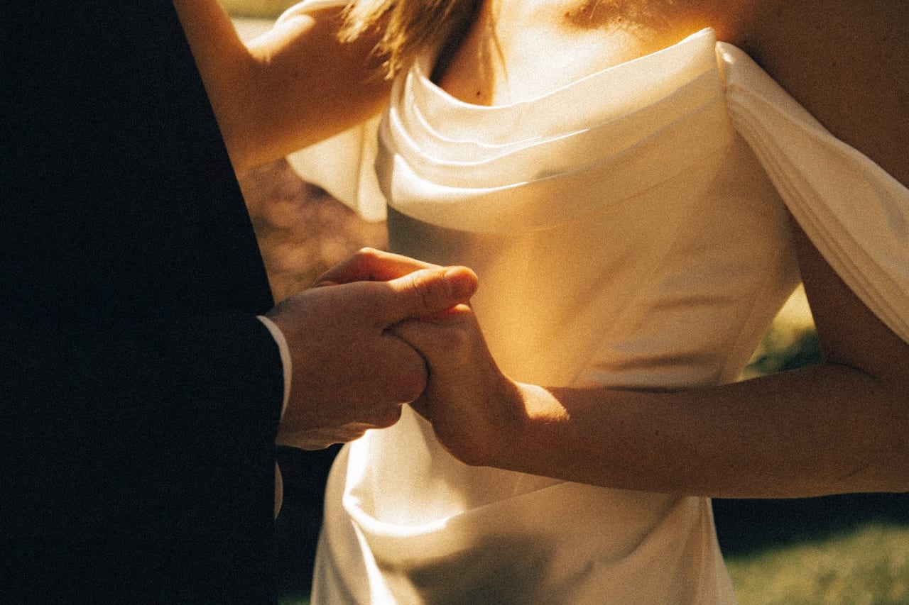 Close-up of a couple holding hands, the bride’s satin gown catching warm light