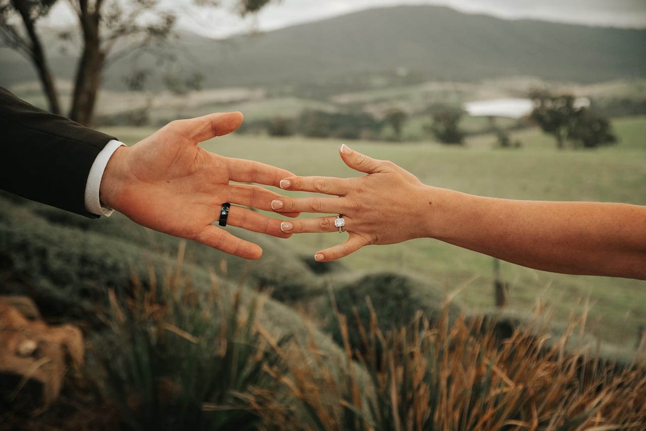 Two hands with wedding rings reaching towards each other over green hills