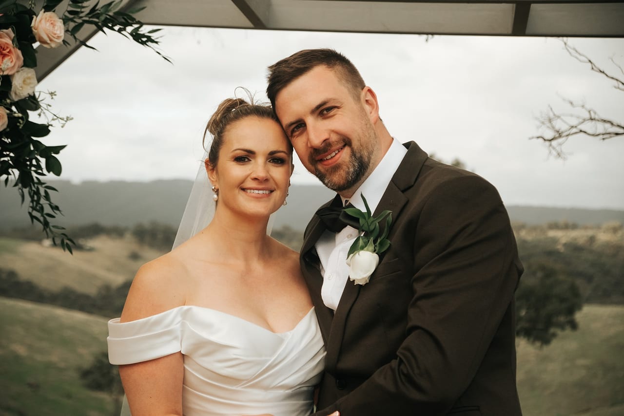 A smiling bride and groom under a canopy with hills behind them