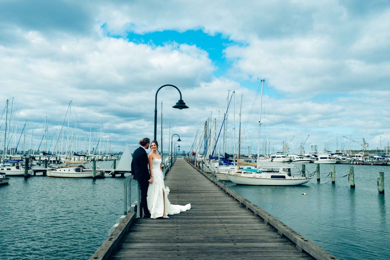 A couple at the end of a long timber pier in a marina under a big sky