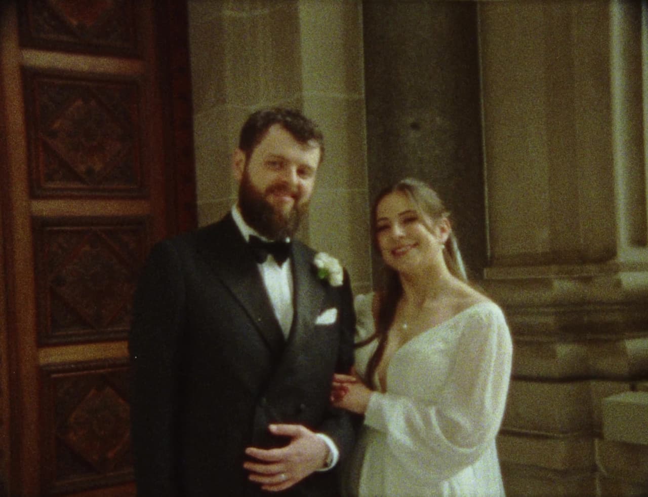 Bianca and Aidan arm in arm in front of a carved wooden door, smiling at the camera