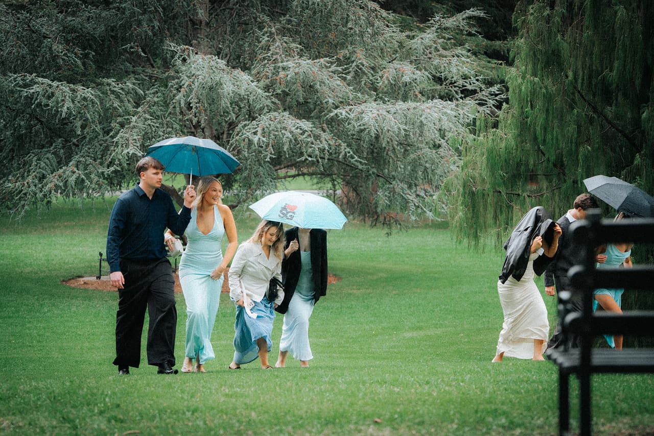 Guests with umbrellas crossing the lawn towards the ceremony, a big cedar and weeping trees behind them