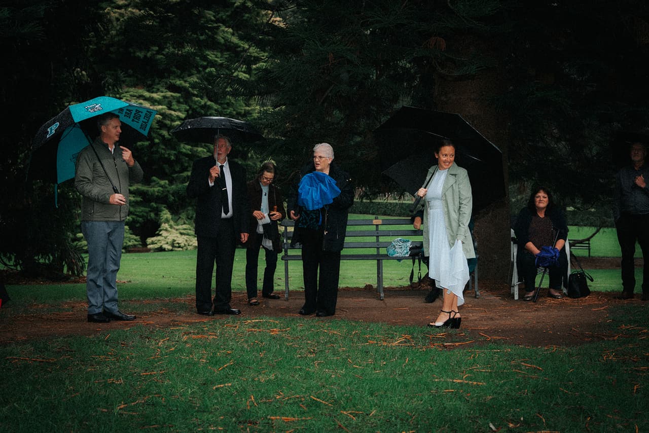Guests waiting with umbrellas beside a garden bench under the pines before the ceremony