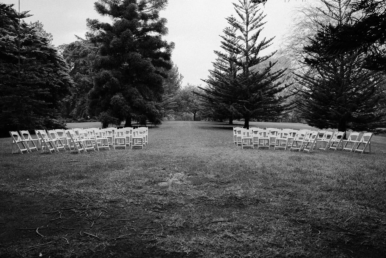Black and white photo of two blocks of white folding chairs on the lawn among the tall pines, ready for the ceremony
