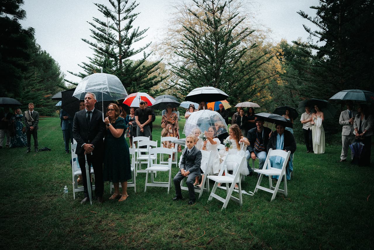 Guests seated and standing under umbrellas among the white chairs during the ceremony, tall pines behind them