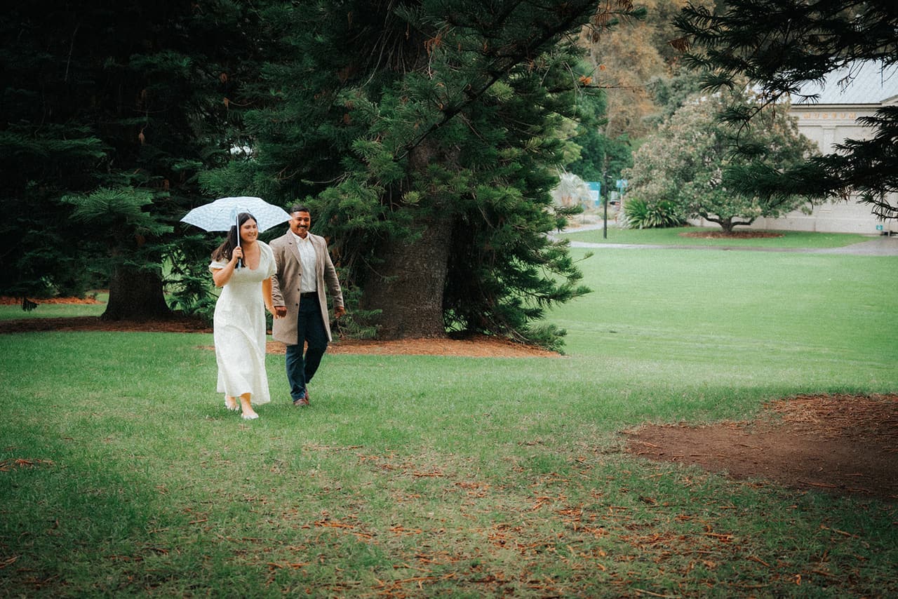 Two guests sharing a white umbrella as they walk across the lawn, a pale heritage building through the trees