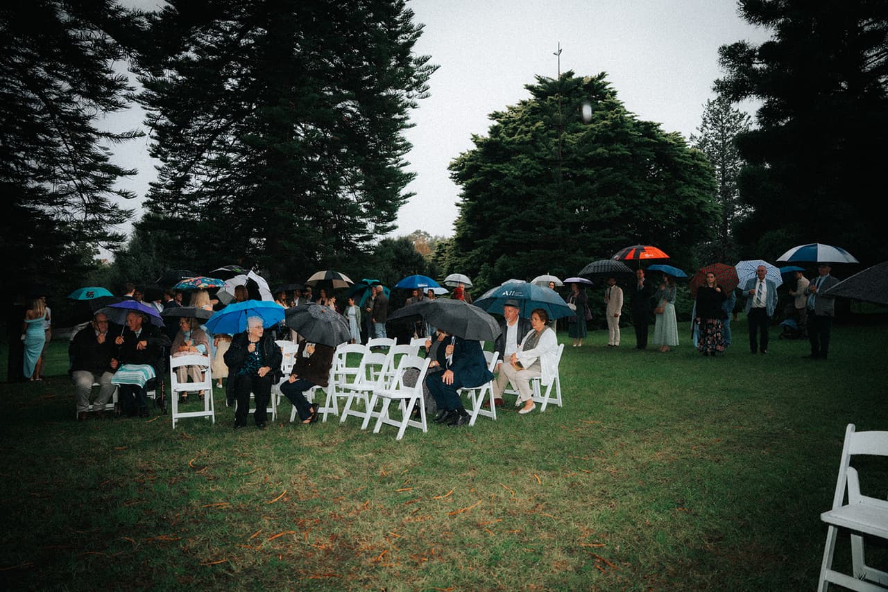 The ceremony clearing from further back, guests under umbrellas in the white chairs and the pines rising behind