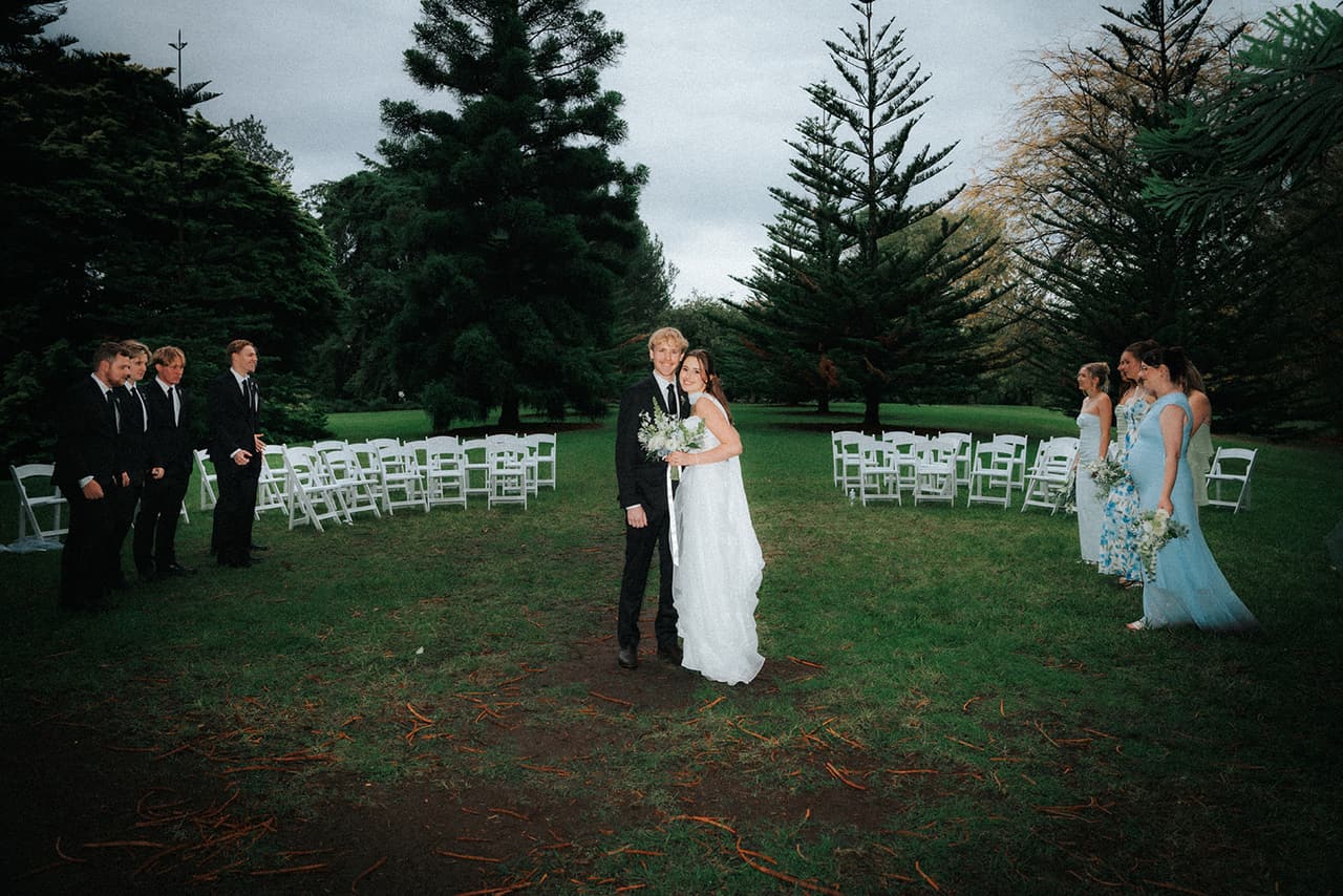 Eli and Charlotte in the middle of the clearing after the ceremony, groomsmen and bridesmaids walking off to either side past the white chairs