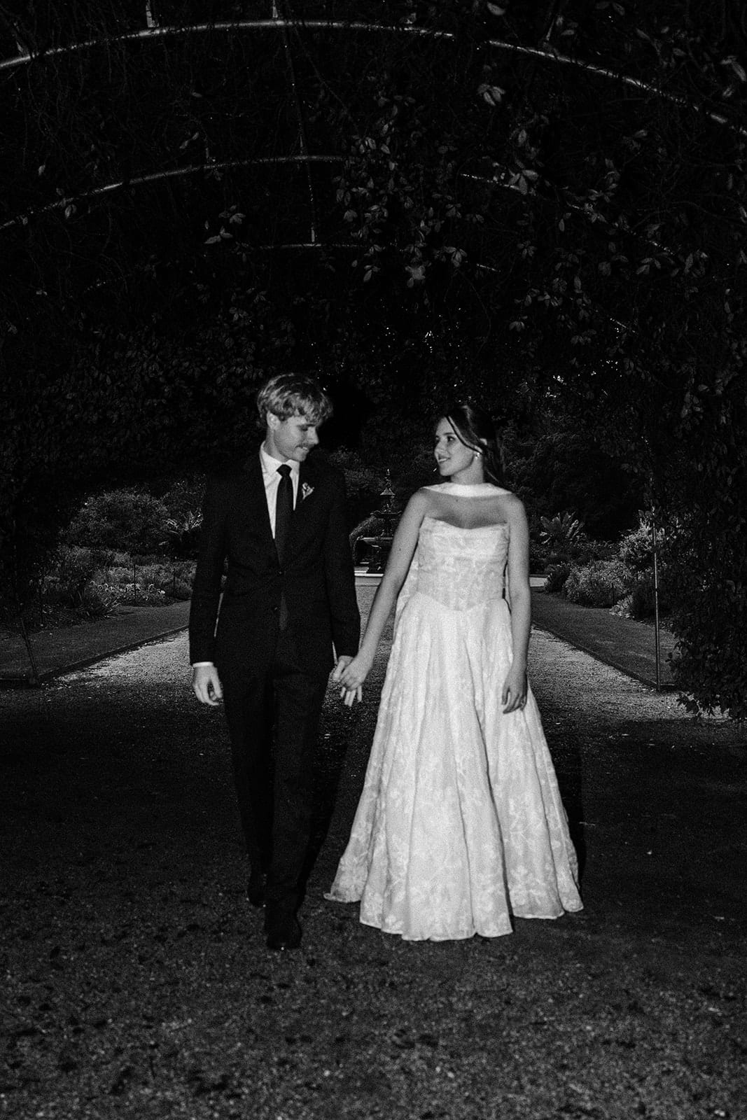 Black and white photo of Eli and Charlotte walking hand in hand along a garden path under an arch of trees