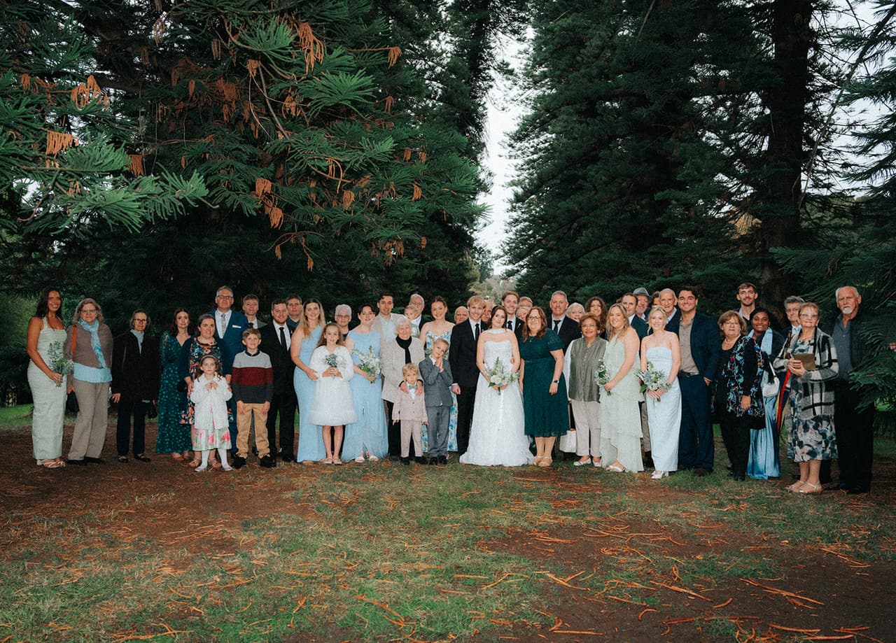 Family and friends gathered around Eli and Charlotte for a group photo beneath the pines
