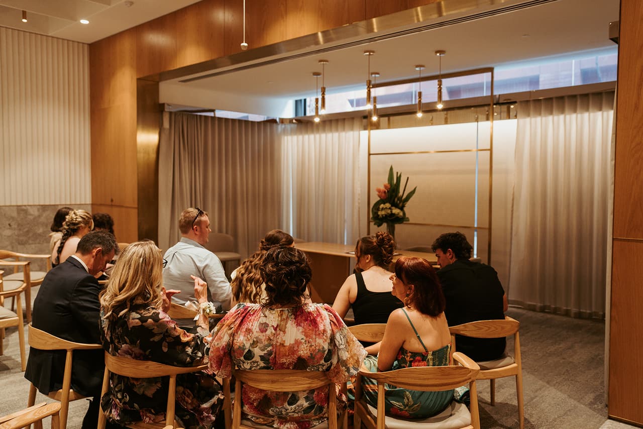 Guests in their seats waiting for the ceremony, a tall vase of flowers on the table in the curtained alcove