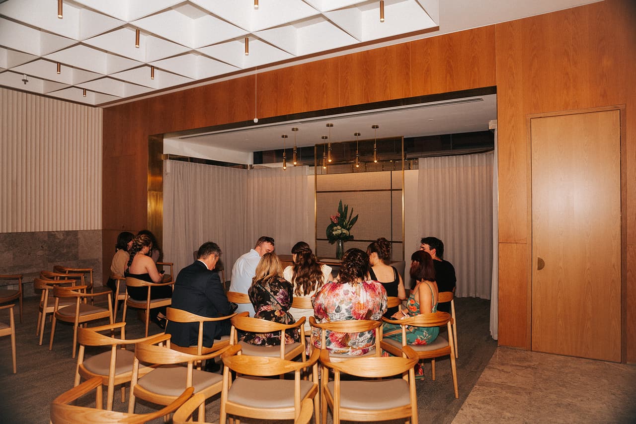 The marriage room from the back corner: timber-panelled walls, rows of timber chairs and the curtained alcove with the table and flowers