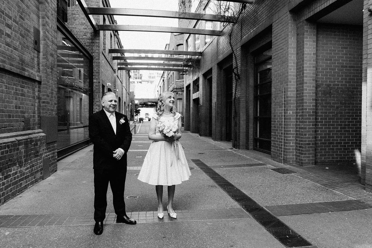 Black and white photo of Jon and Terrie standing in the middle of the laneway outside the registry, the glass canopy overhead