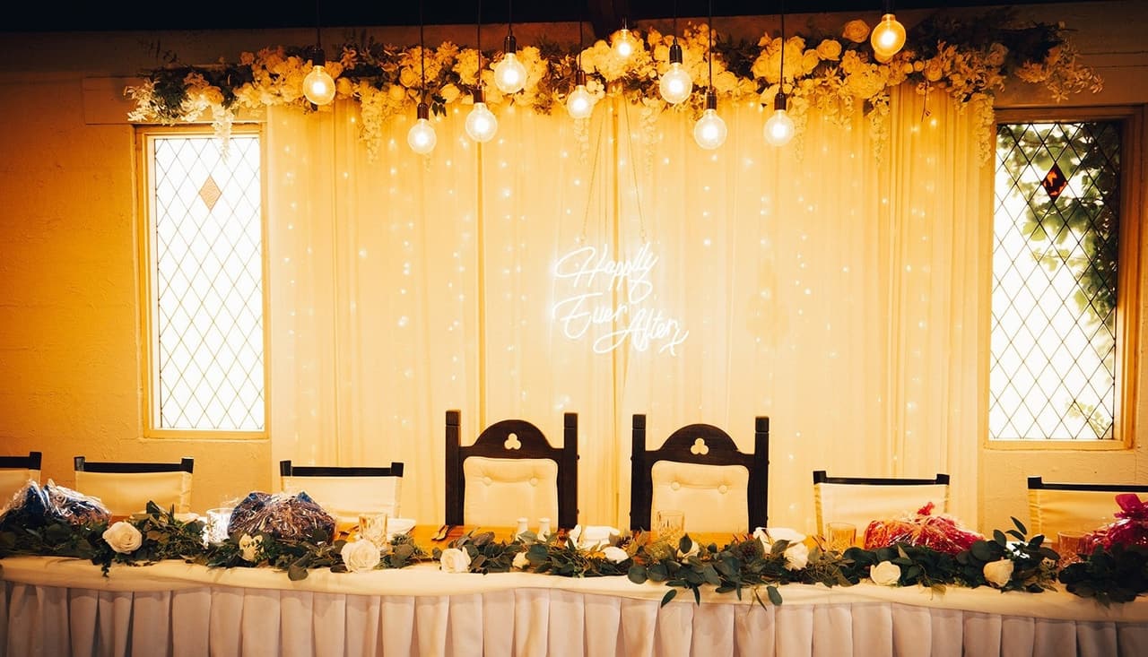 The bridal table in front of a curtain of fairy lights, hanging bulbs, flowers and a neon sign