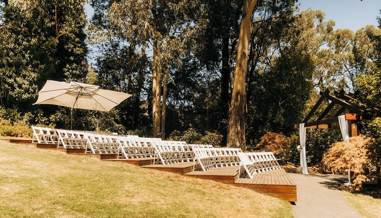 The ceremony site before the guests arrive: tiers of timber decking with white chairs set into the lawn, a market umbrella and tall gums