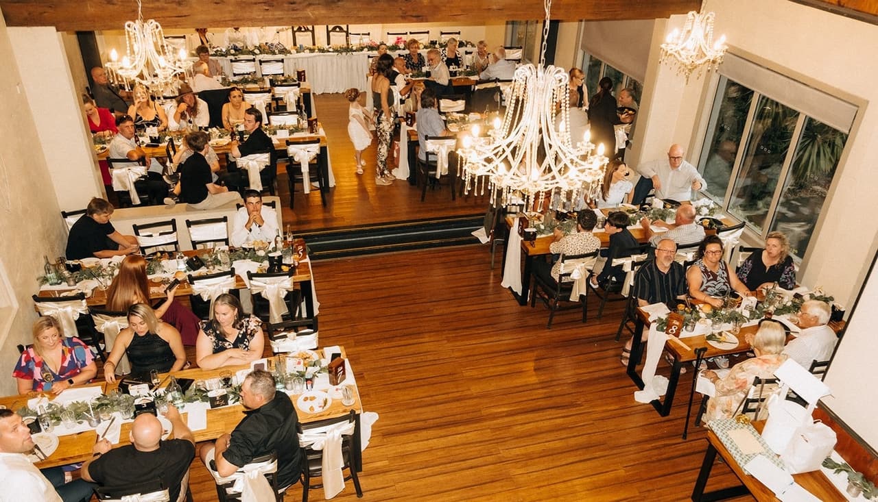 The reception hall from the mezzanine during dinner, guests at long timber tables under chandeliers