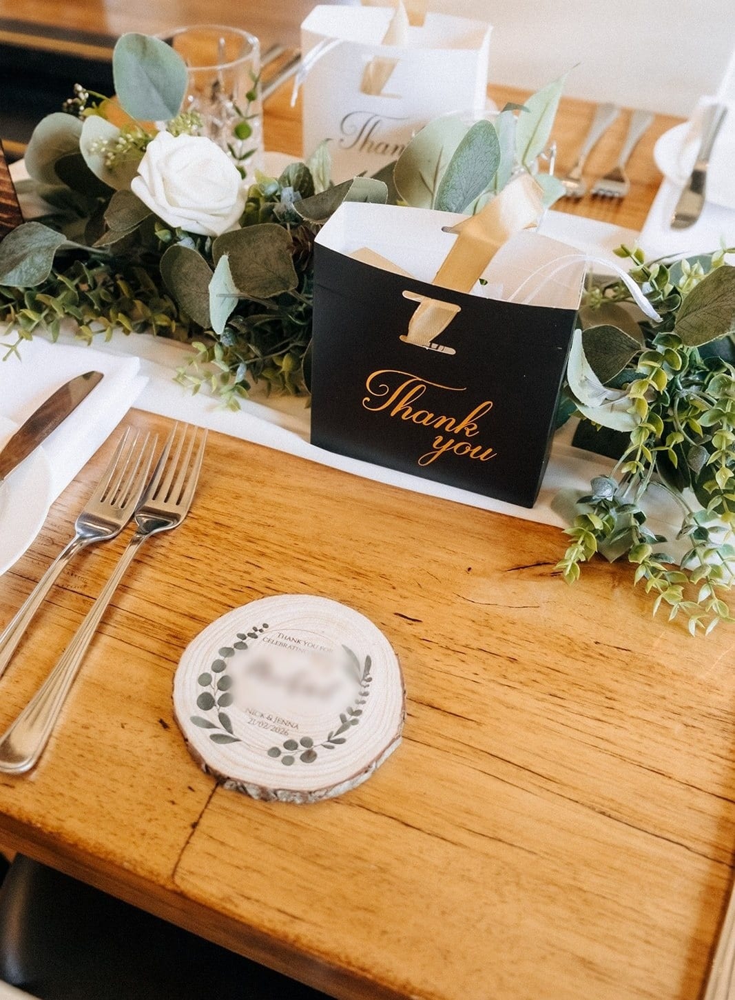 A place setting on a timber table with a wooden coaster and a black thank-you bag