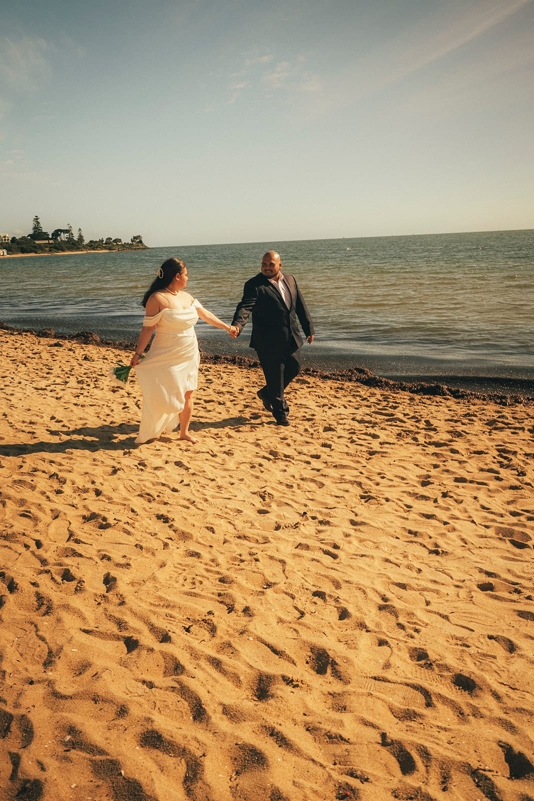 Martina and Diego walking hand in hand across the sand at Brighton Beach, the bay and a wooded headland behind them