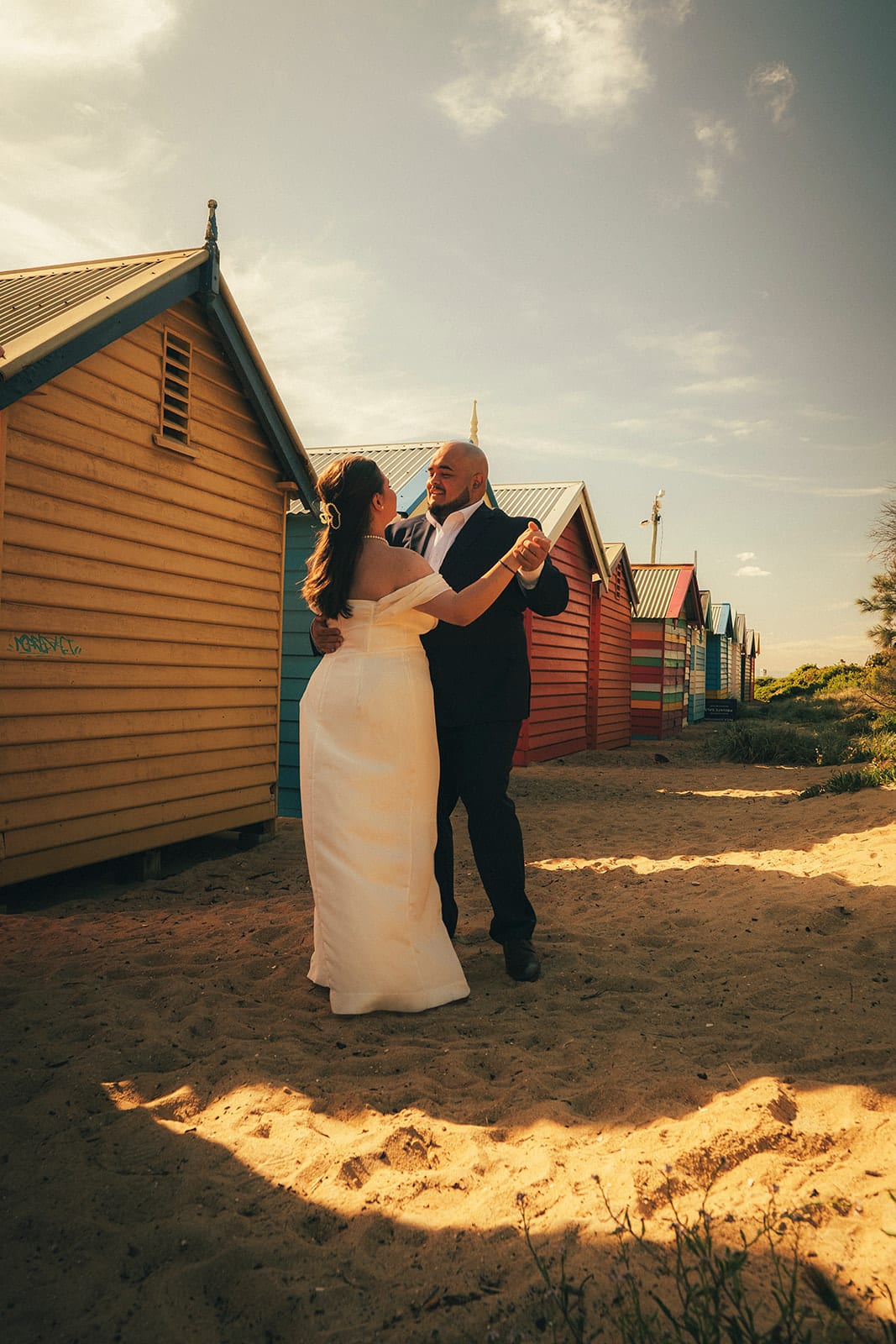Martina and Diego dancing on the sand in front of a yellow bathing box, the row of colourful boxes behind them