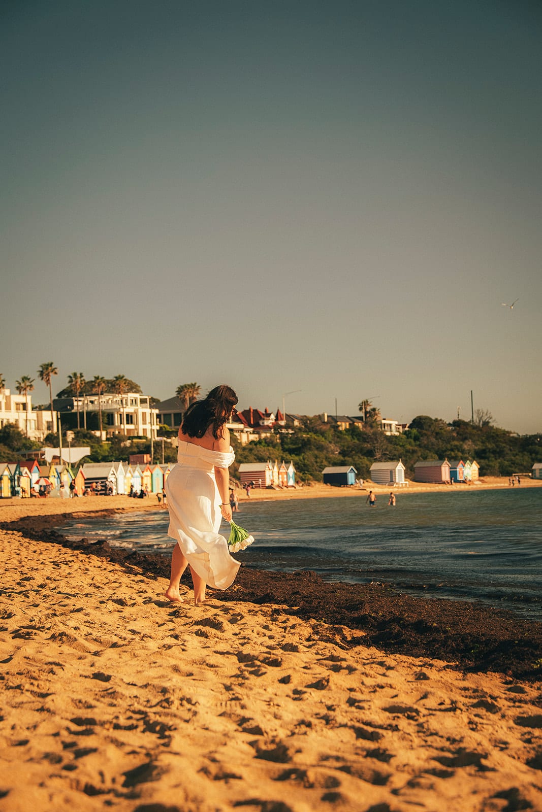 Martina walking at the water’s edge, the bathing boxes strung along the curve of the beach and houses on the rise above