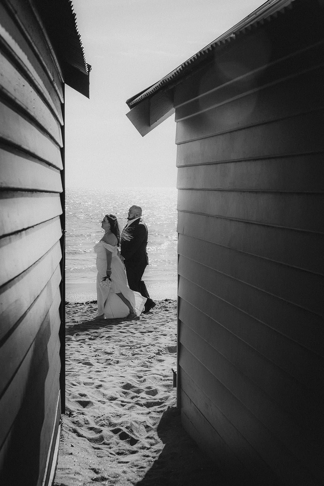 Black and white photo of Martina and Diego on the sand by the water, seen through the narrow gap between two bathing boxes