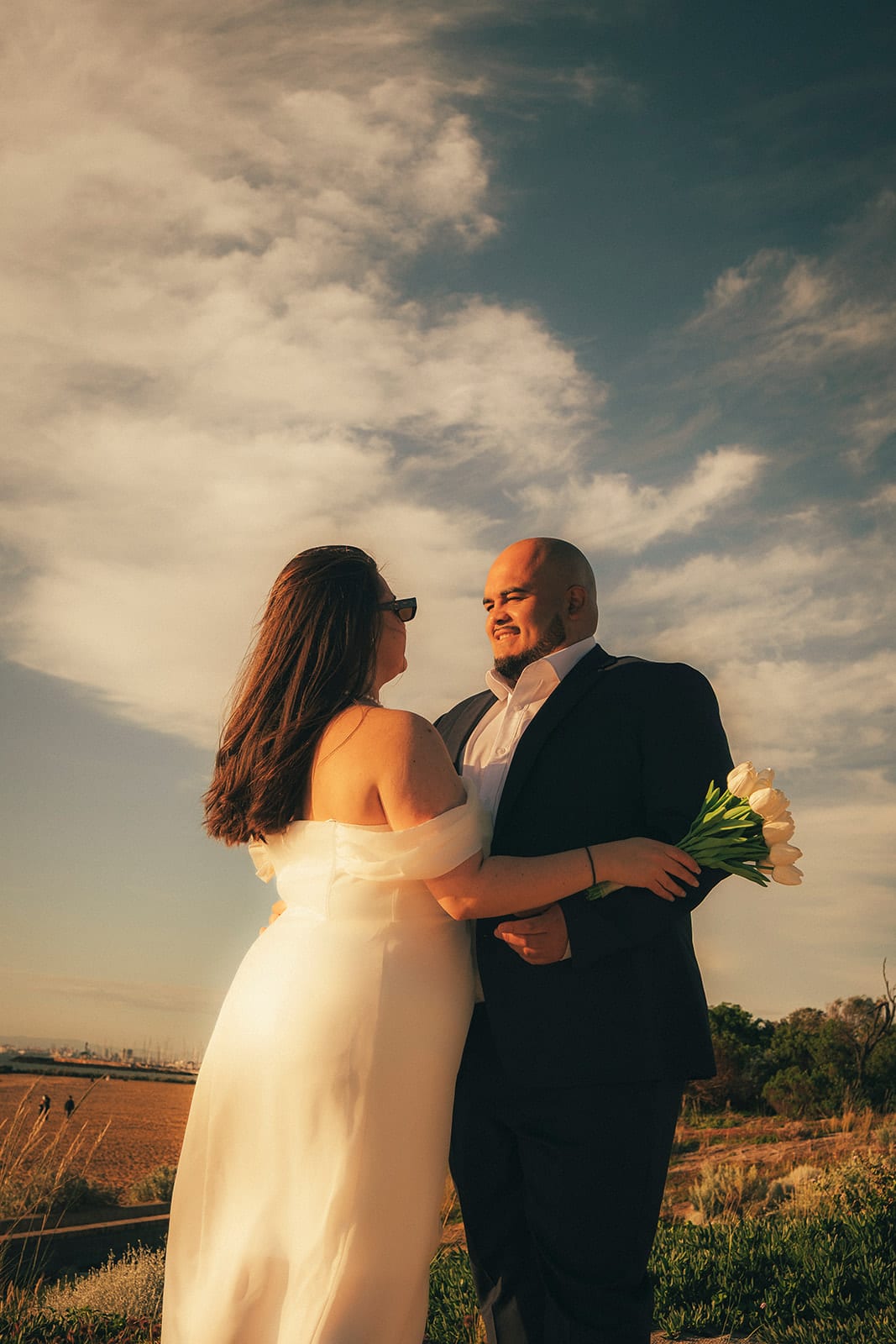 Martina and Diego holding each other on the grassy foreshore above the beach, clouds over the bay