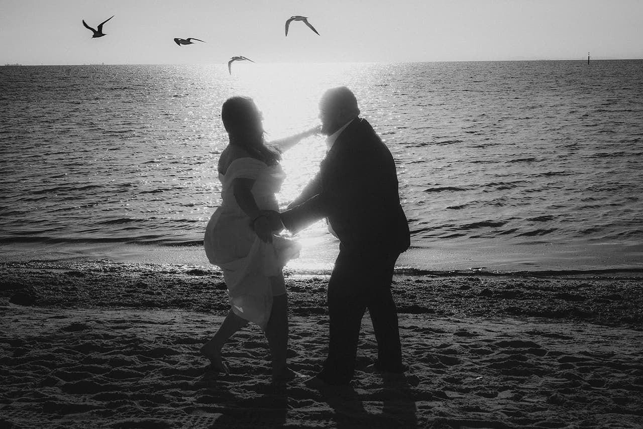 Black and white photo of Martina and Diego in silhouette by the water as seagulls fly overhead