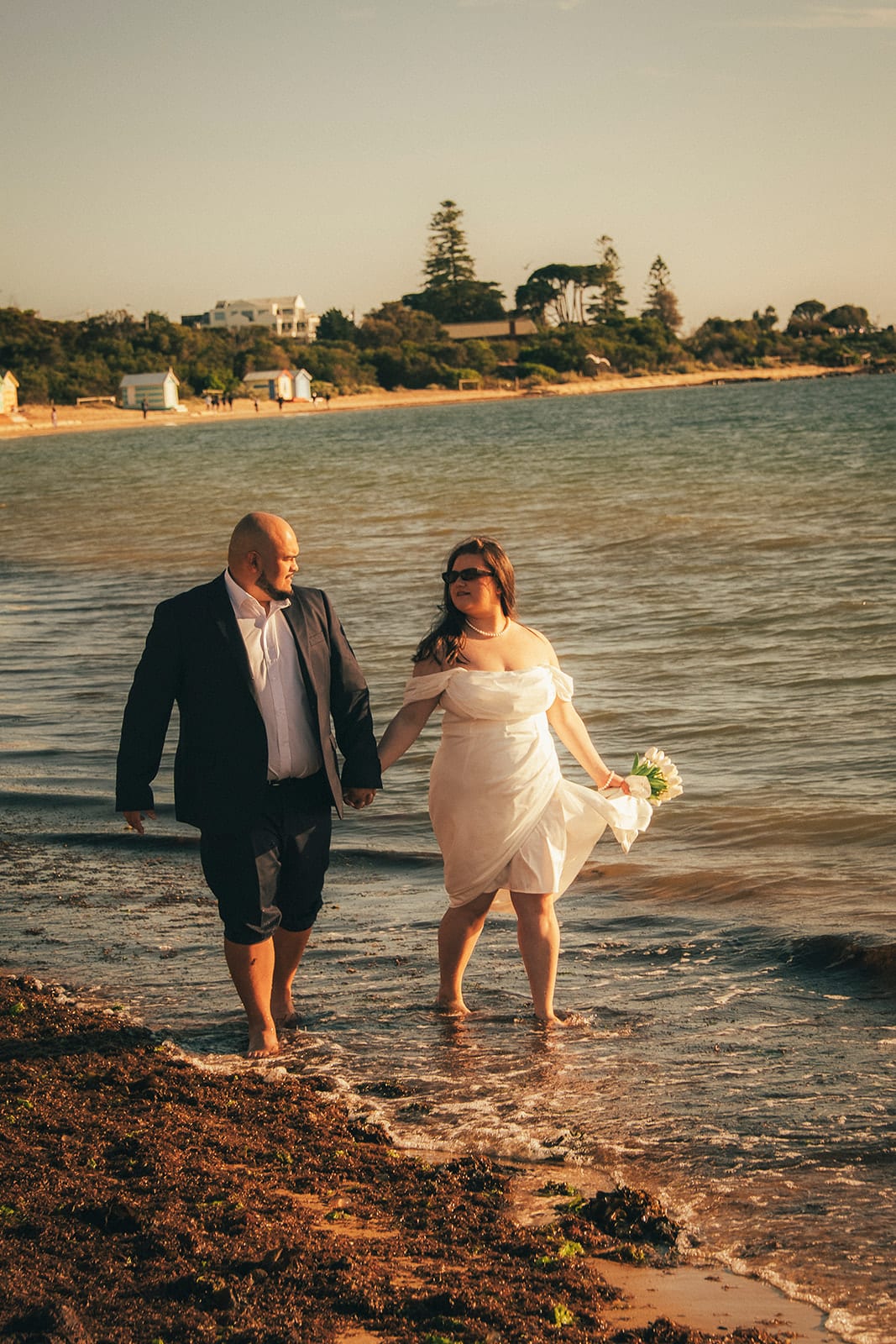 Martina and Diego wading hand in hand in the shallows, a wooded headland and houses across the water
