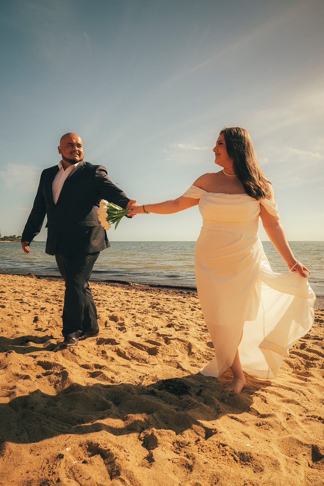 Martina and Diego walking hand in hand along the sand by the water, her dress swinging