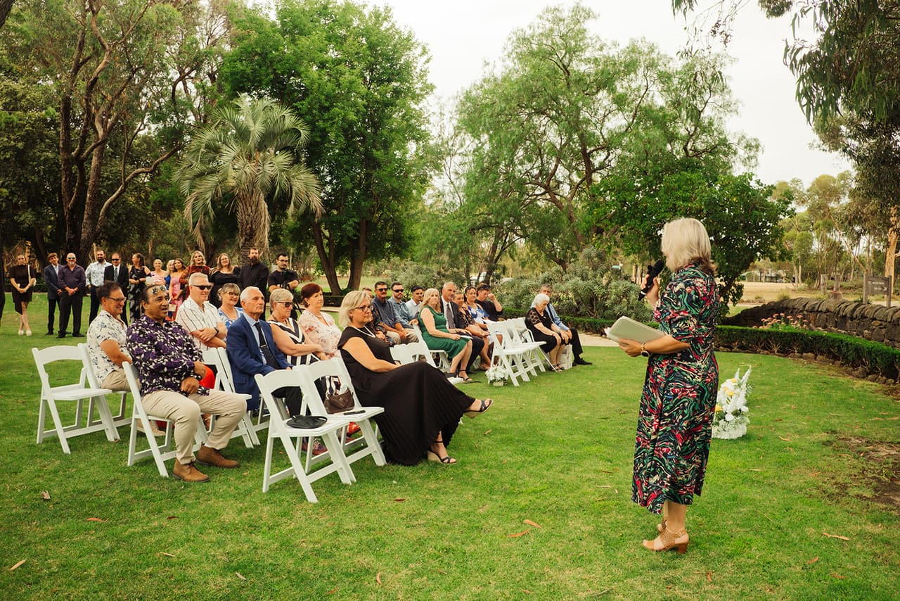 The celebrant welcoming the seated guests, white folding chairs set out on the lawn under the trees
