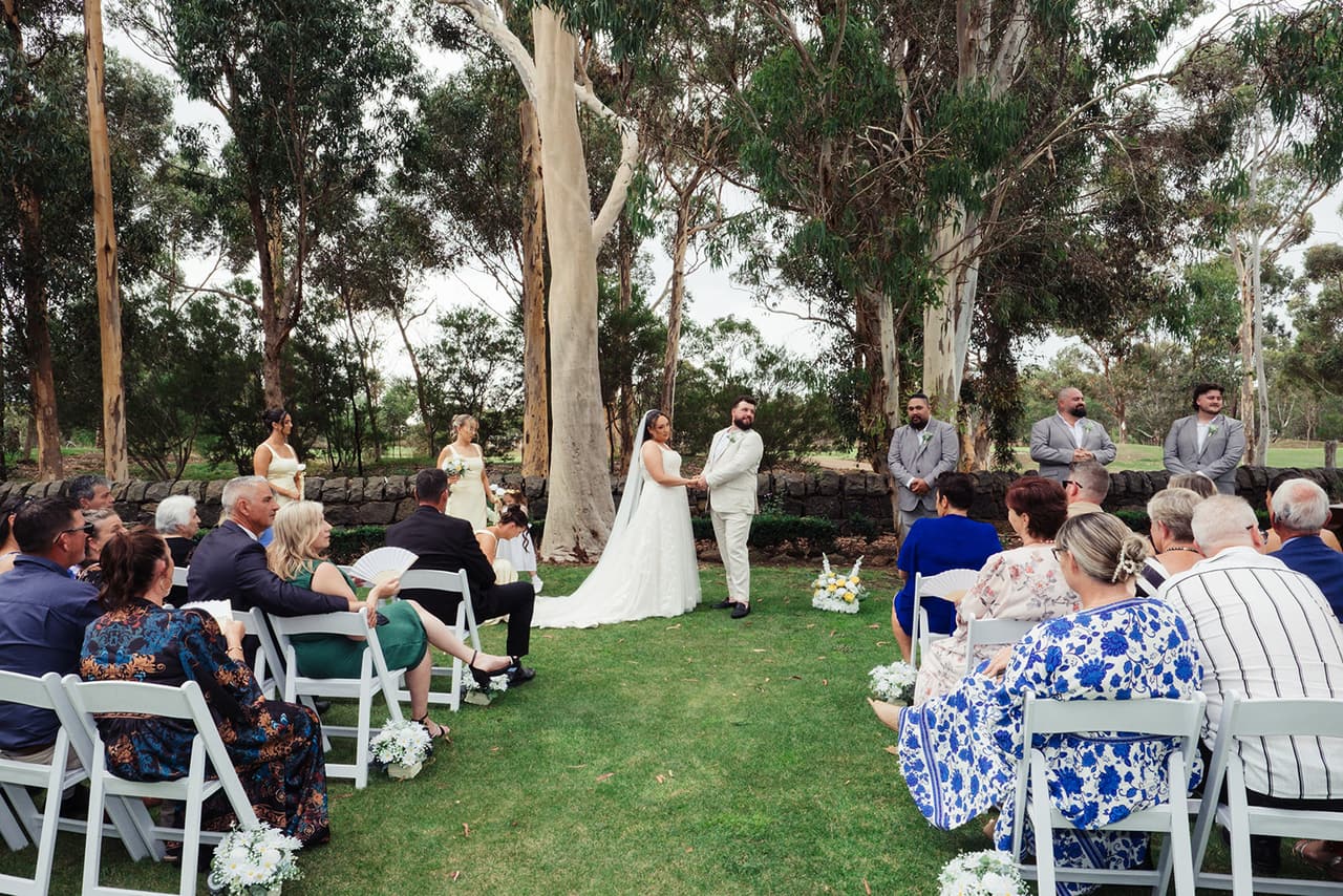 The ceremony from the back of the aisle, Karina and Bronson facing each other under the tall gums by the old stone wall
