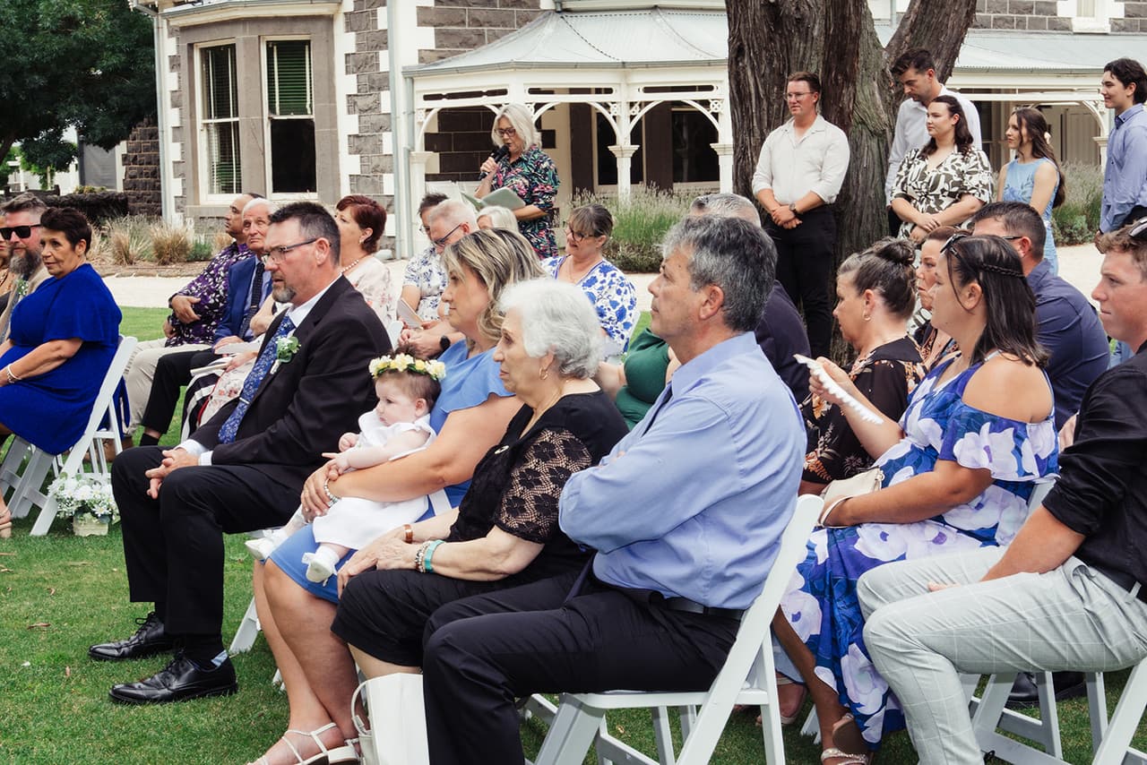 Guests seated in white chairs for the ceremony, the bluestone homestead and its veranda behind them