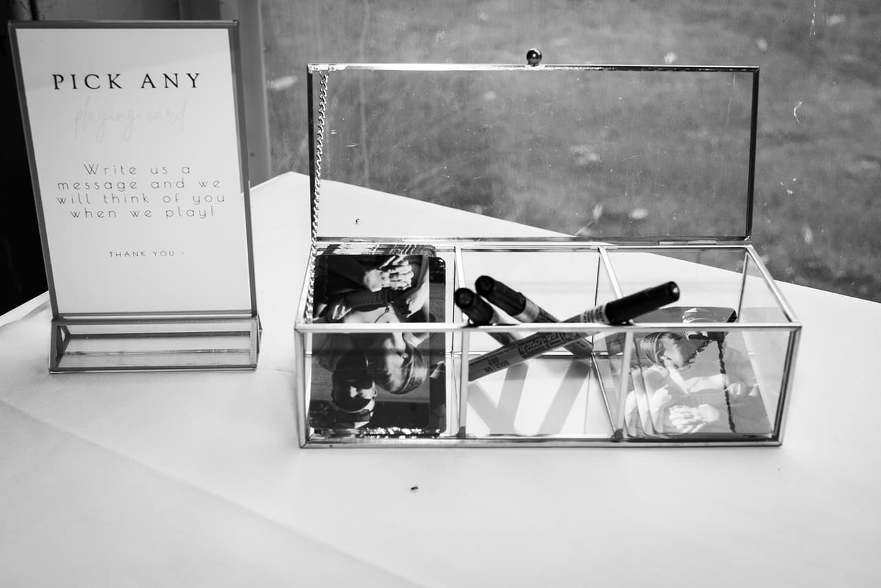 Black and white photo of a glass box of playing cards and pens beside a sign asking guests to write a message