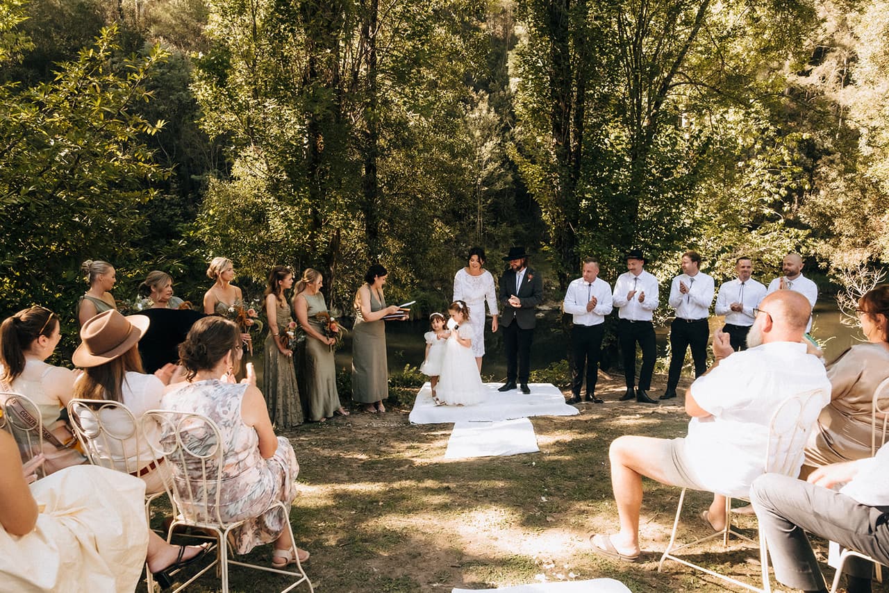 The ceremony from behind the guests, Lana and Todd at the front between the bridesmaids and groomsmen, the river trees beyond