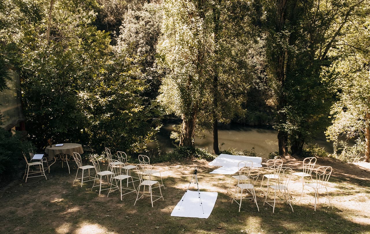 White wire chairs and a white runner set out under the trees by the river before the ceremony