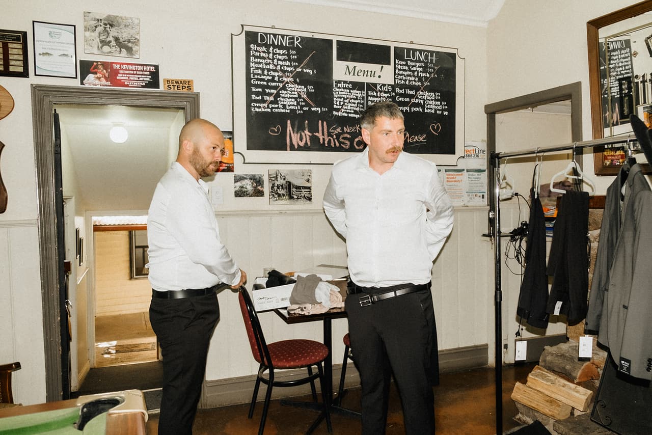 Two groomsmen dressing in the room off the bar, the hotel’s chalkboard menus on the wall and their suits on a rack