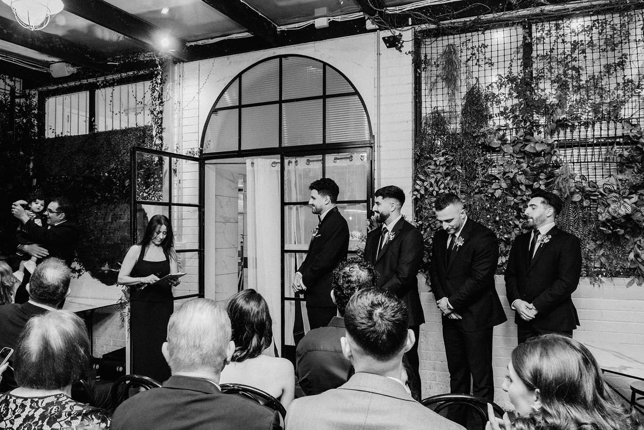 Black and white photo of the courtyard from behind the guests, the groomsmen and celebrant waiting under the arched window