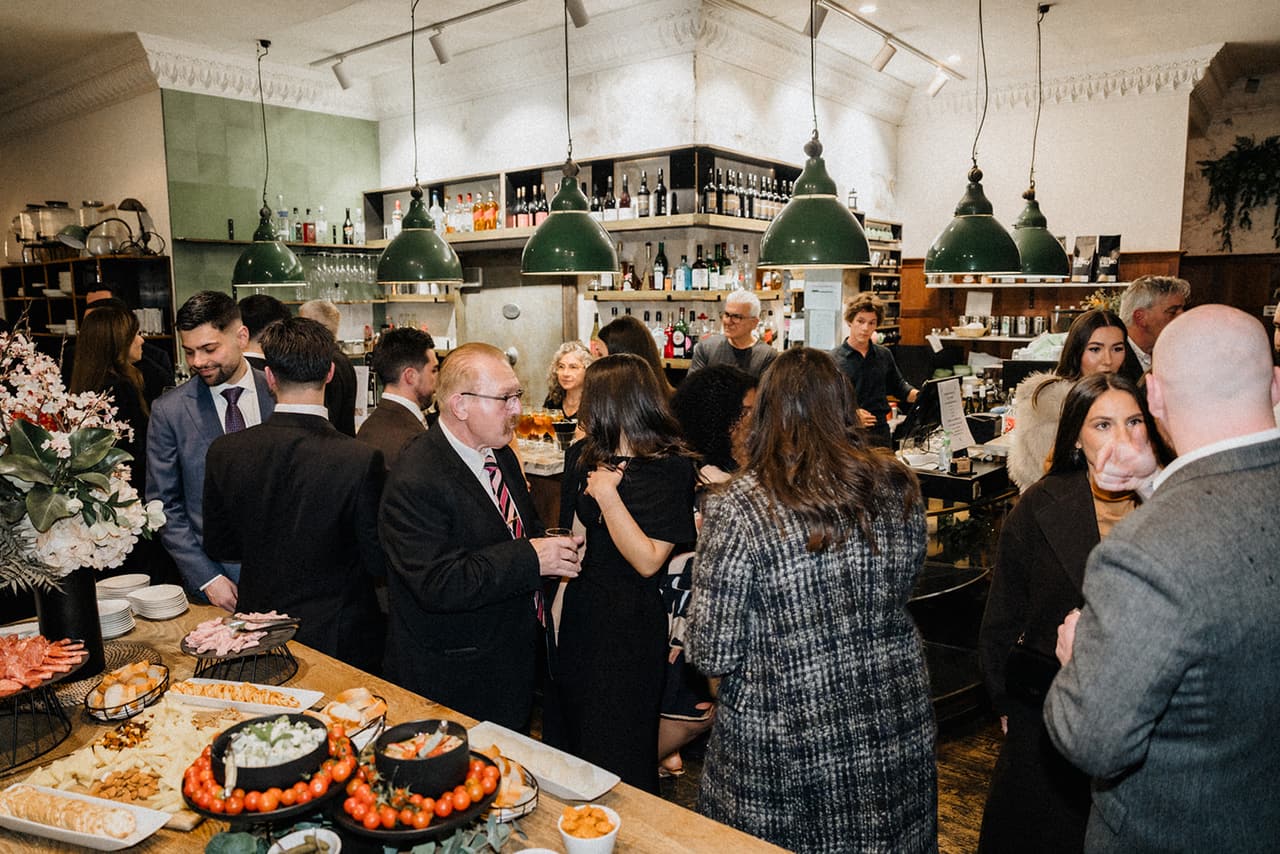 Guests around the grazing table in front of the bar, green enamel lamps hanging over shelves of bottles
