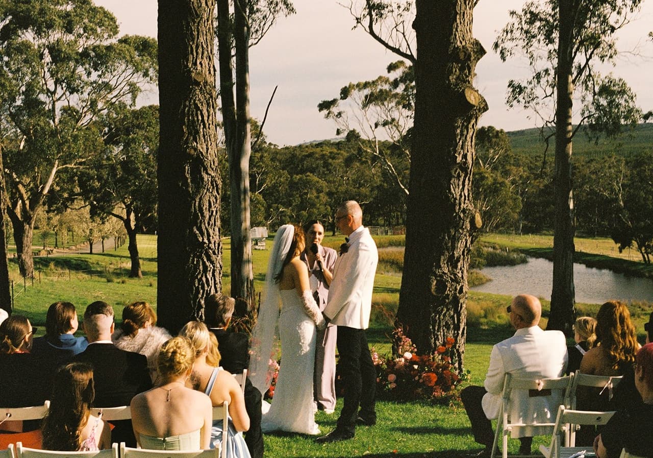 The ceremony from behind the guests, the couple under tall trees with a dam and paddocks beyond, on 35mm film